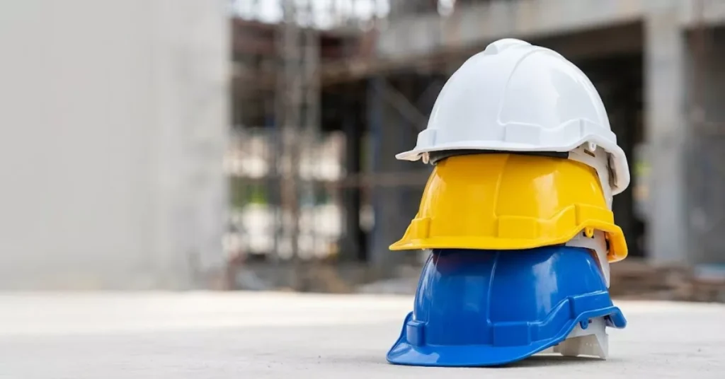 Tumpukan helm proyek warnna biru, kuning, dan putih di sebuah site konstruksi.
